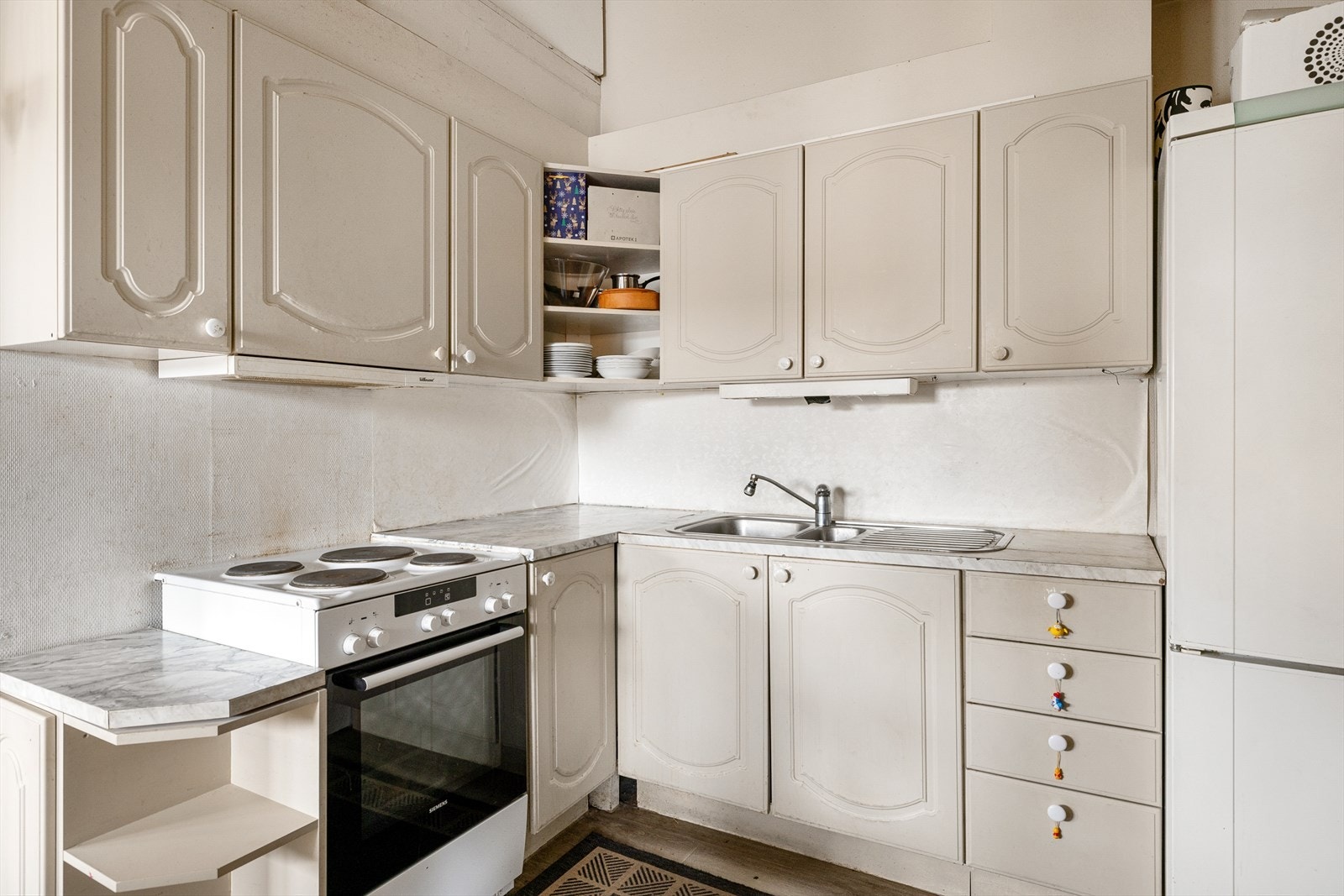 Innredningen består av lyse, profilerte fronter, benkeplate i laminat med nedfelt kum i rustfritt stål, belysning under overskap og tapet på vegg over benkeskap. Frittstående komfyr og kjøl/fryseskap. Kjøkkenventilator med avtrekk ut. Galleribilde