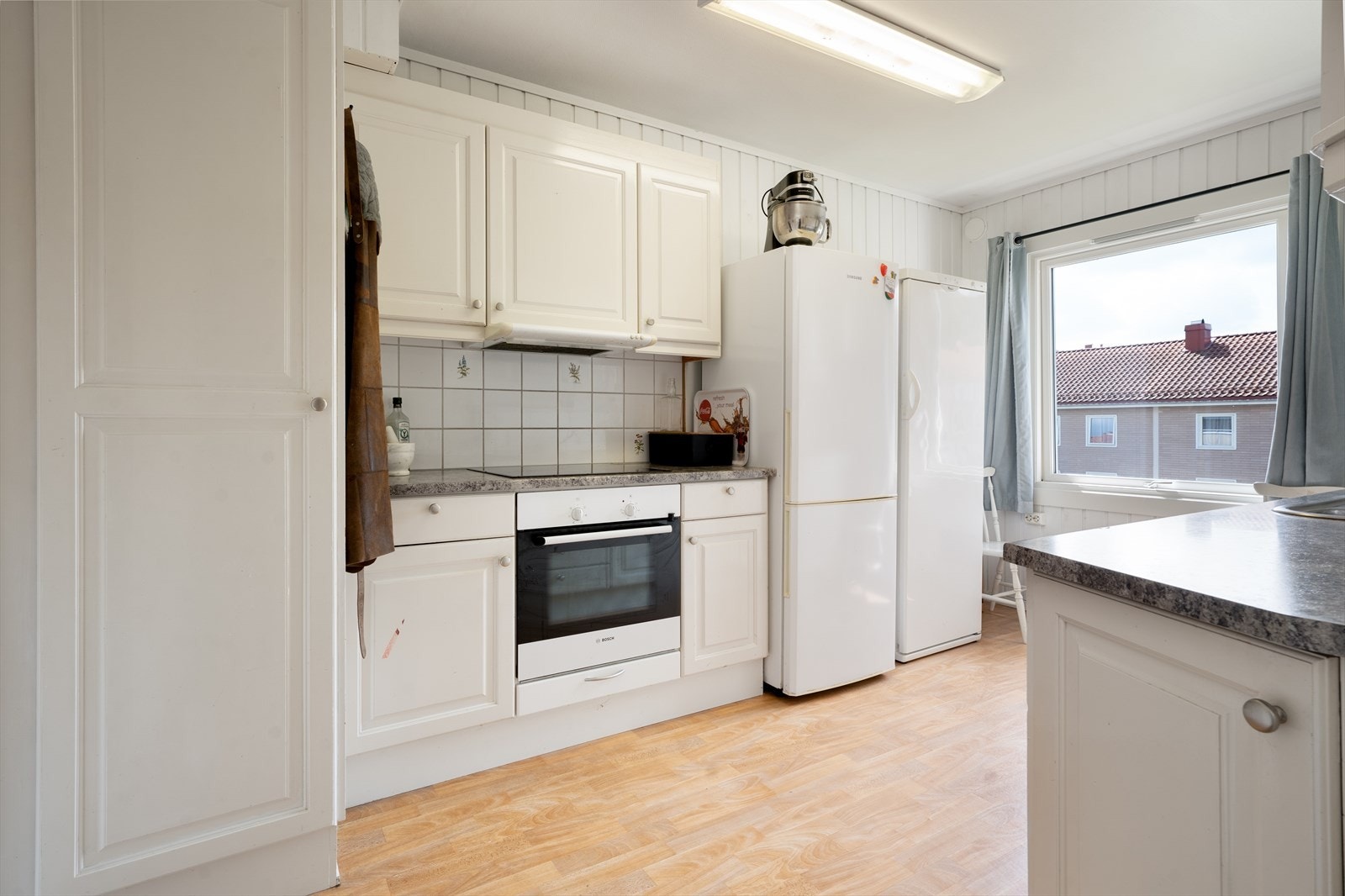 Kjøkkenet har en hvit innredning med profilerte fronter, laminat benkeplate med oppvaskkum i rustfritt stål og flis over kjøkkenbenk. Her er det også belysning og el-stikk. Galleribilde