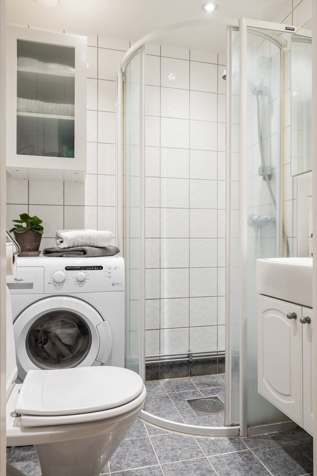 Badet består av veggskap med speilfronter over servant. Dusj med faste felt og skyvedører i herdet glass, servant i helstøpt plate, gulvstående toalett og opplegg for vaskemaskin. Galleribilde