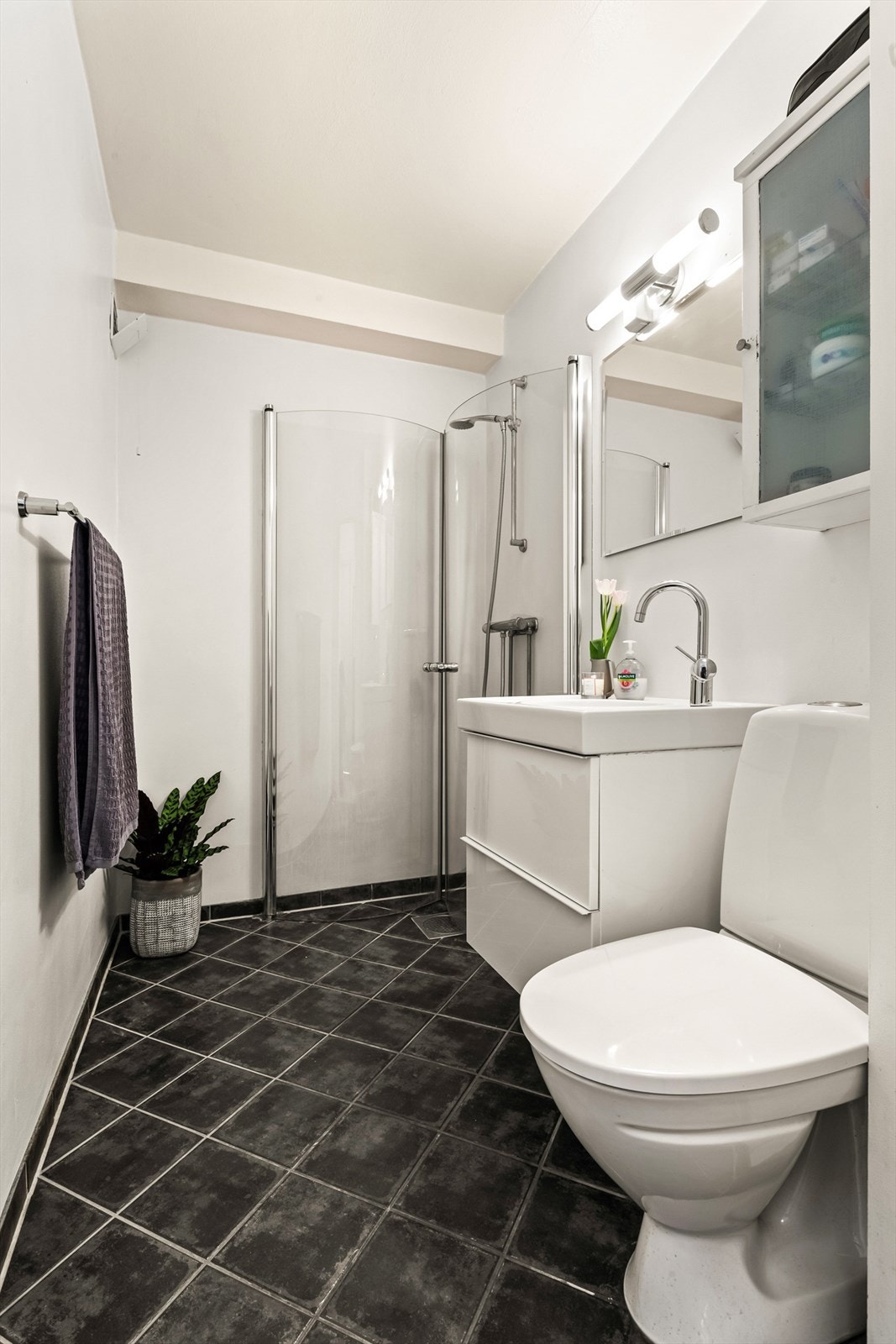 Eldre baderom utstyrt med gulvstående klosett, dusjhjørne med innfellbare dører og en servant med tilhørende innredning. Galleribilde