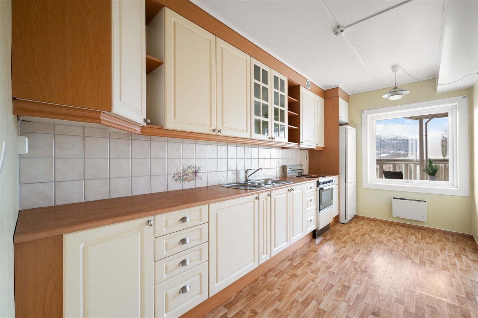 Kjøkkenet er målt til 10,5m² og inneholder profilerte fronter, et overskap med glassfronter, laminat benkeplate med fliser over, stål oppvaskkum og ventilator. Galleribilde