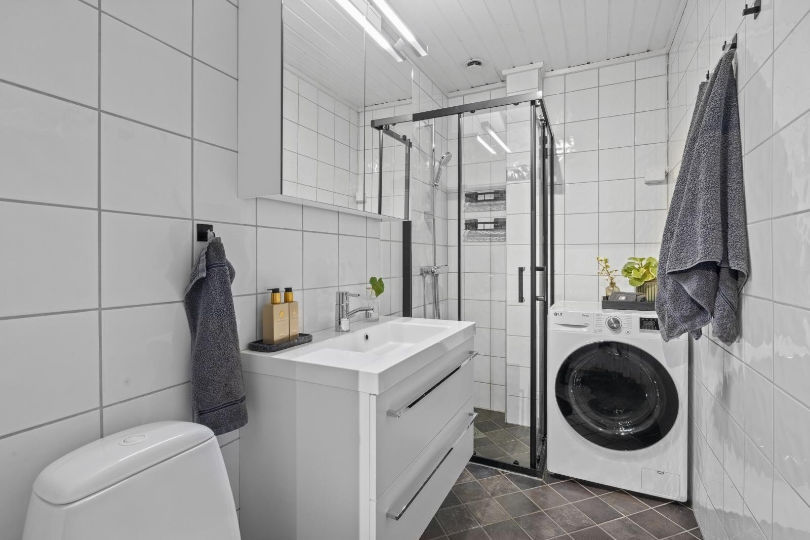 Servant med skuffeseksjon, speilskap, gulvmontert klosett, dusjhjørne med skyvedører og opplegg for vaskemaskin. Galleribilde