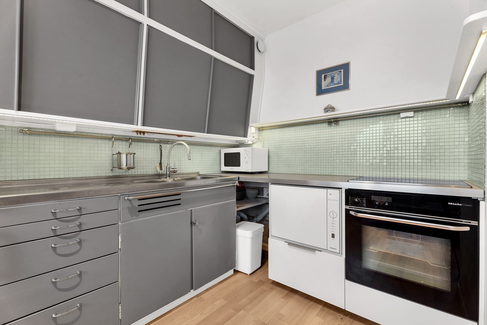 Original kjøkkeninnredning med malte slette fronter, laminat benkeplate med glassmosaikkfliser, dobbel oppvaskkum med helt benkebeslag, opplegg for oppvaskmaskin, integrert stekeovn og koketopp. Galleribilde