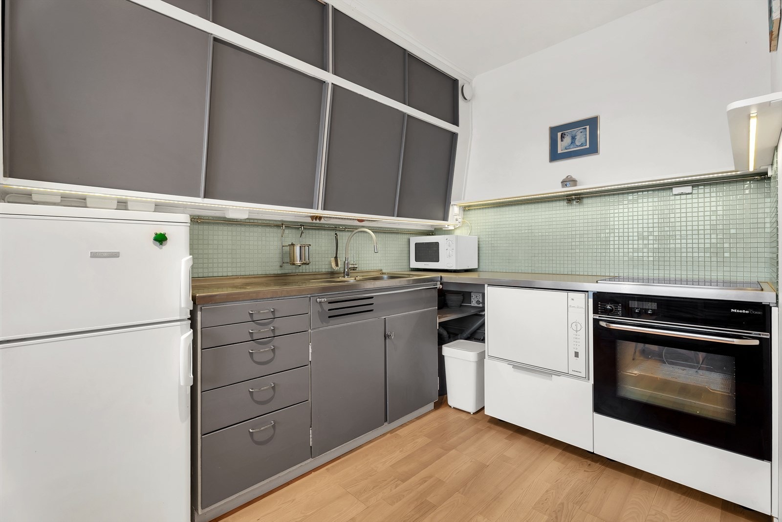 Original kjøkkeninnredning med malte slette fronter, laminat benkeplate med glassmosaikkfliser, dobbel oppvaskkum med helt benkebeslag, opplegg for oppvaskmaskin, integrert stekeovn og koketopp. Galleribilde