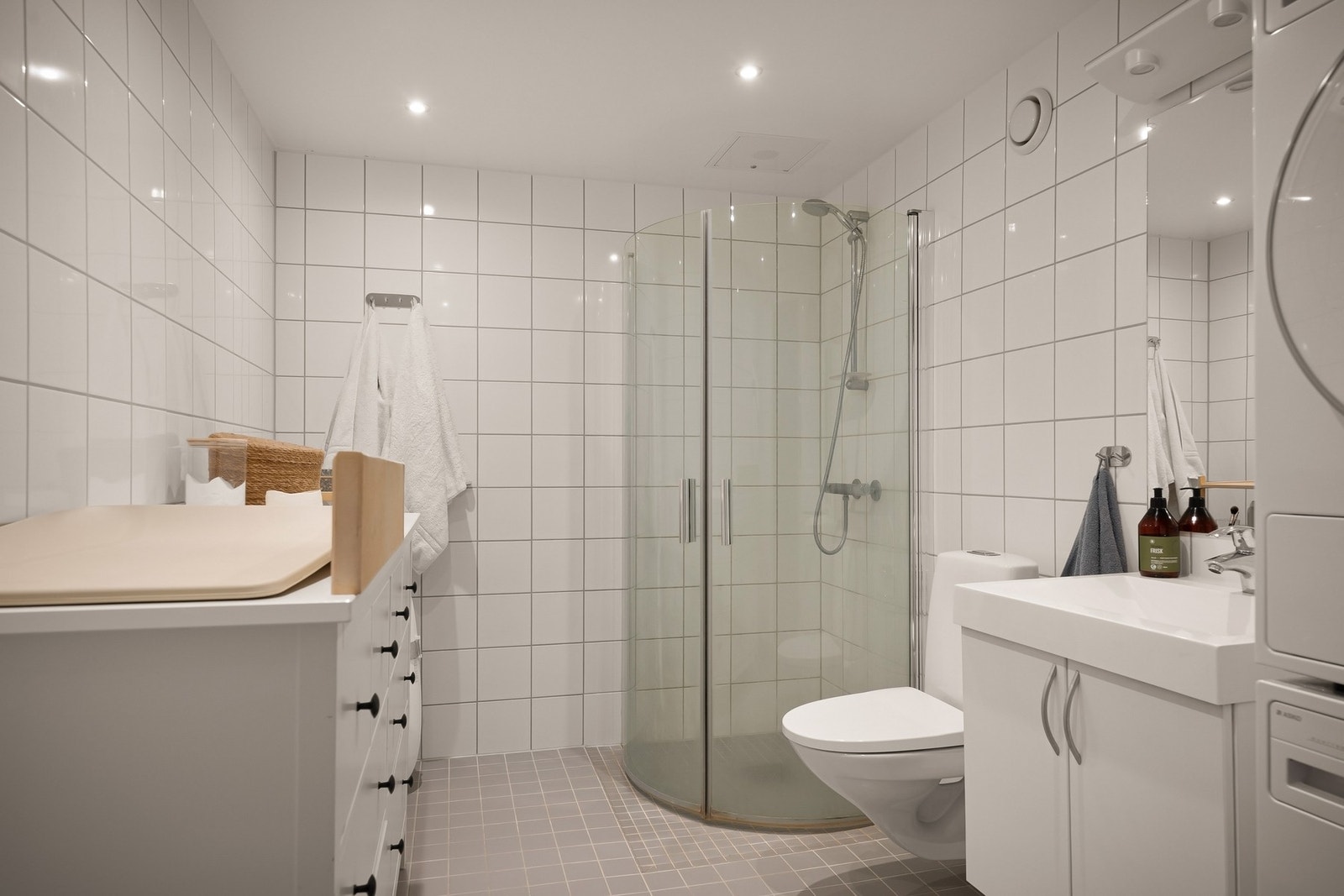 Badet fremstår tidløst og moderne med lyse fliser på vegger og grå fliser på gulv, samt mosaikkfliser i dusjsonen Galleribilde