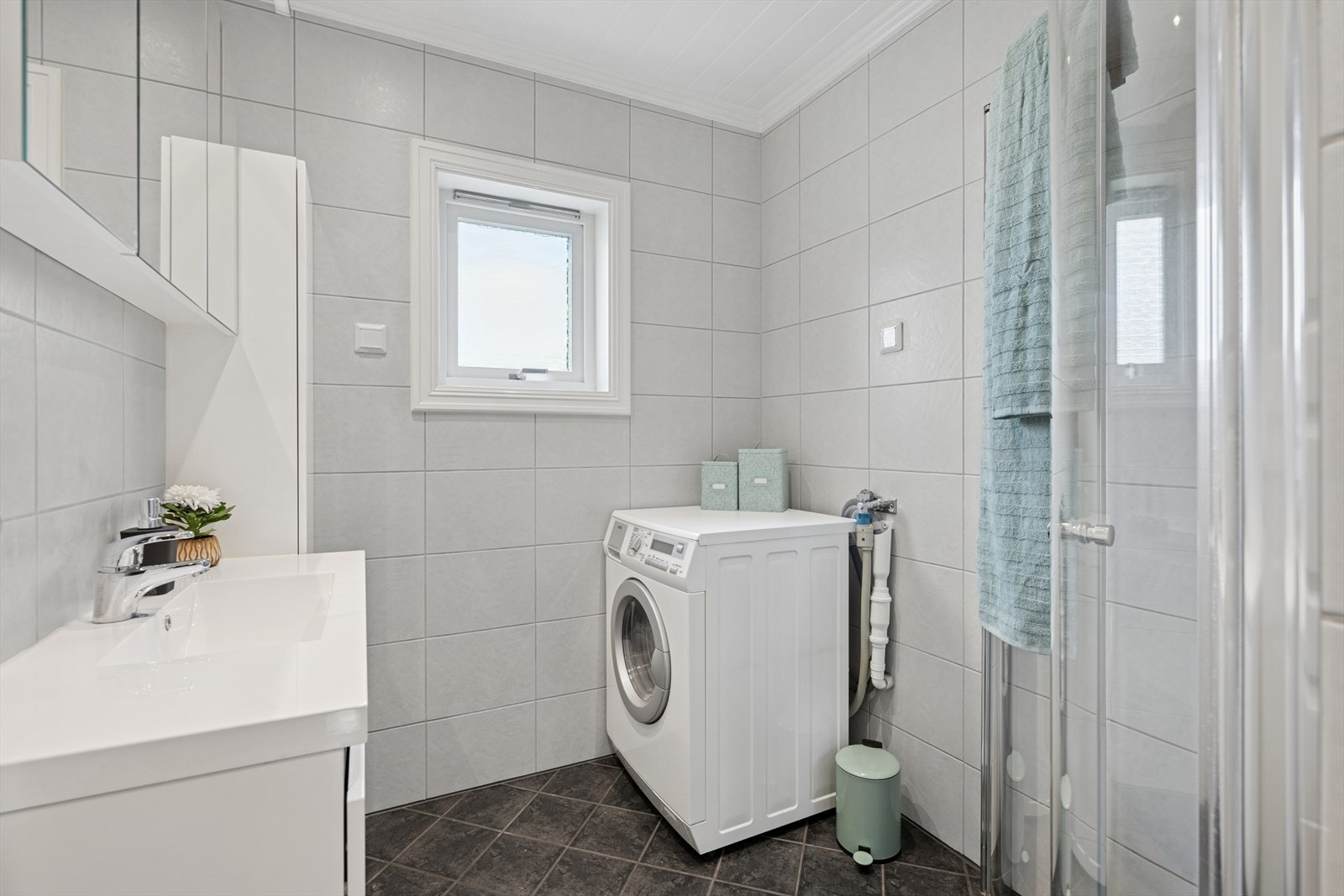 Rommet er innredet med nedfelt servant i moderne baderomsinnredning, samt dusjhjørne med glassvegger som gir god plass og et rent, stilfullt uttrykk. Det er opplegg for vaskemaskin, noe som gjør badet både praktisk og funksjonelt for daglig bruk. Galleribilde