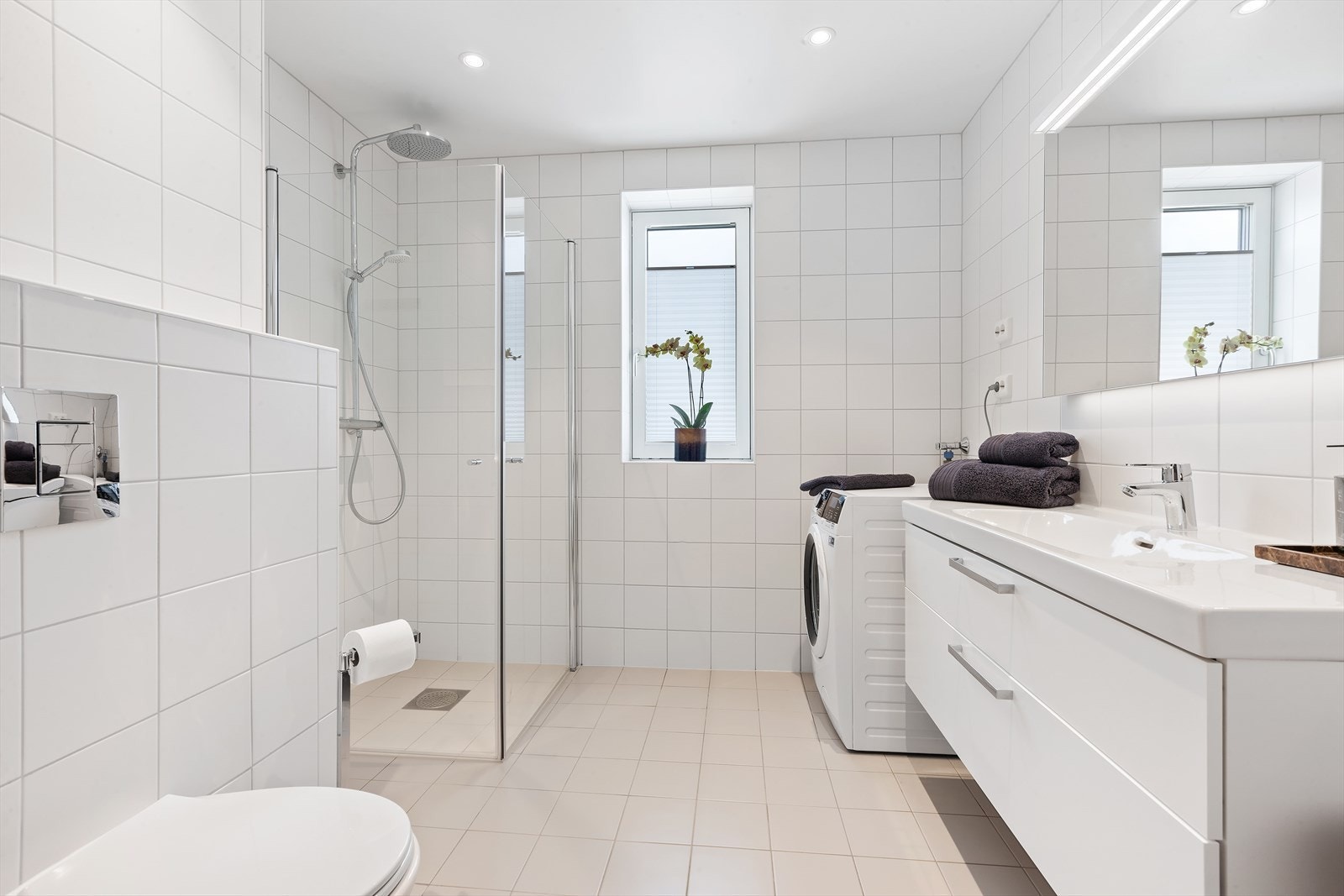 Hovedbadet er av god størrelse og har flislagte overflater på gulv og vegger. Her er det dusjhjørne med glassdører, vegghengt toalett, en pen servantinnredning og opplegg for vaskemaskin. Galleribilde