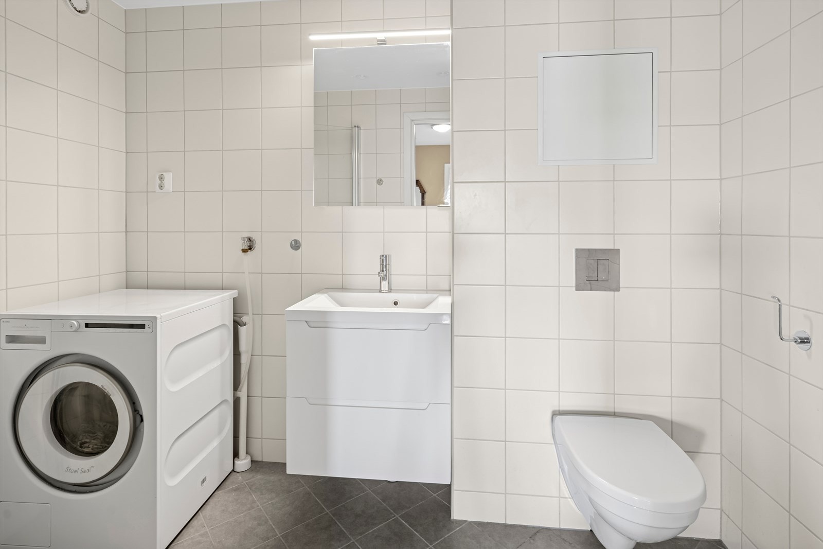 Badet fremstår tidløst og delikat med flislagte overflater, moderne innredning, varmekabler i gulv og opplegg for vaskemaskin. Galleribilde