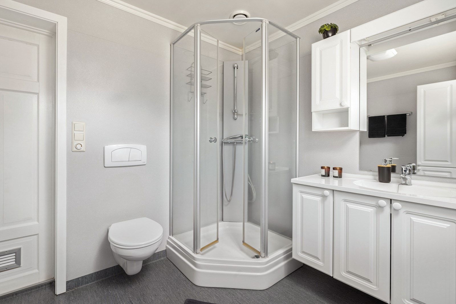 Lys baderomsinnredning med heldekkende servant, speil med belysning og vegghengte skap over, samt ekstra høyskap. Baderommet er også utstyrt med dusjkabinett og vegghengt toalett. Galleribilde