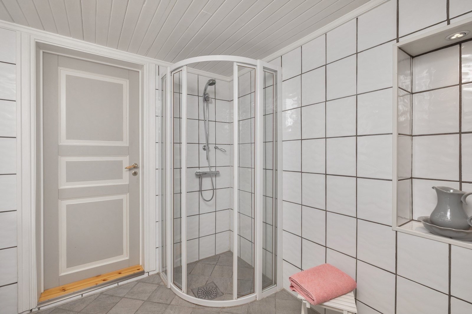 Baderommet har grå fliser på gulv, elektrisk gulvvarme, vegger med hvite fliser kombinert med lysegrå panelplater, samt mekanisk avtrekk. Galleribilde