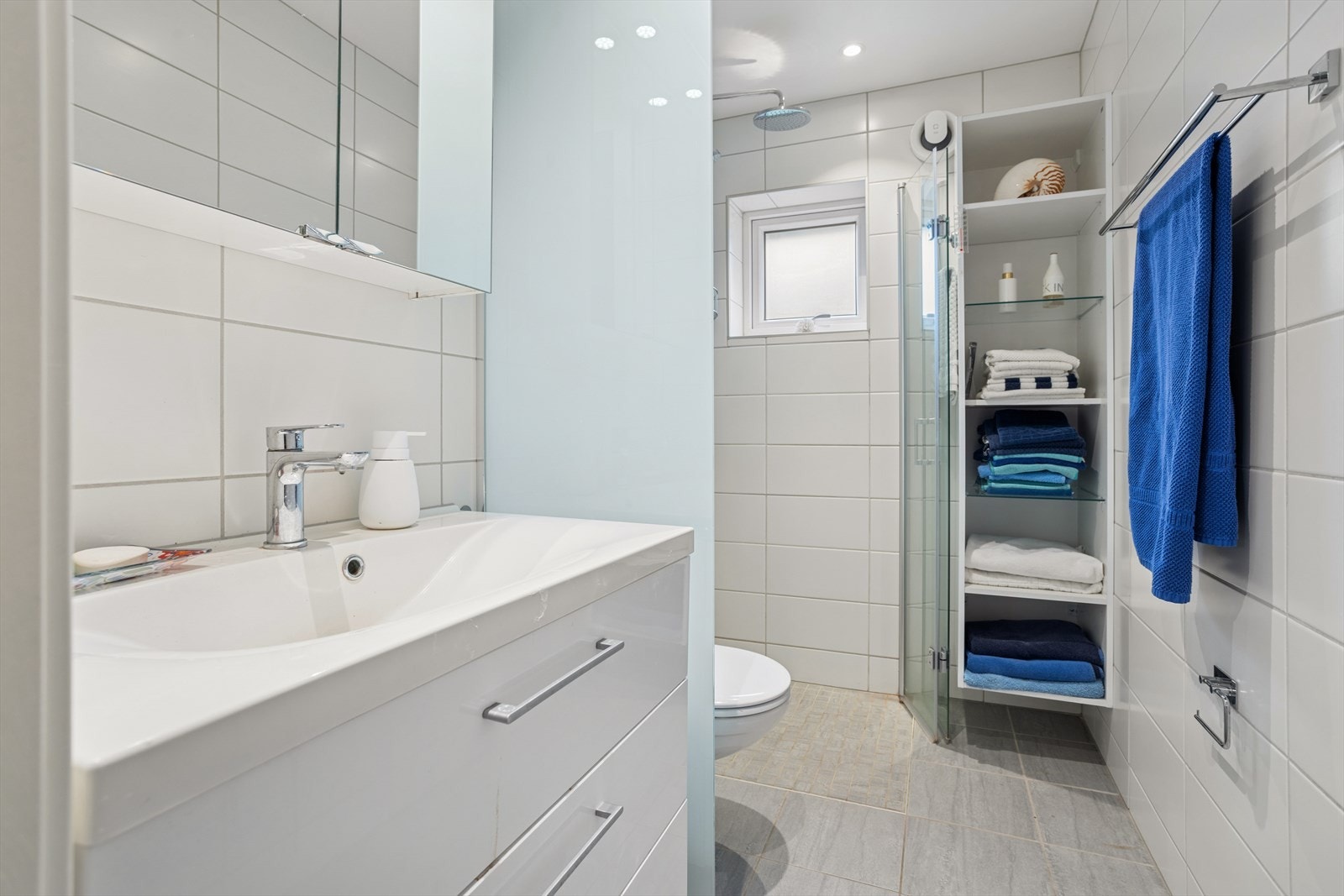 Delikat baderom med fliser på gulv og vegger, innredet med servant med skap, vegghengt toalett, dusjhjørne og god hylleplass. Galleribilde