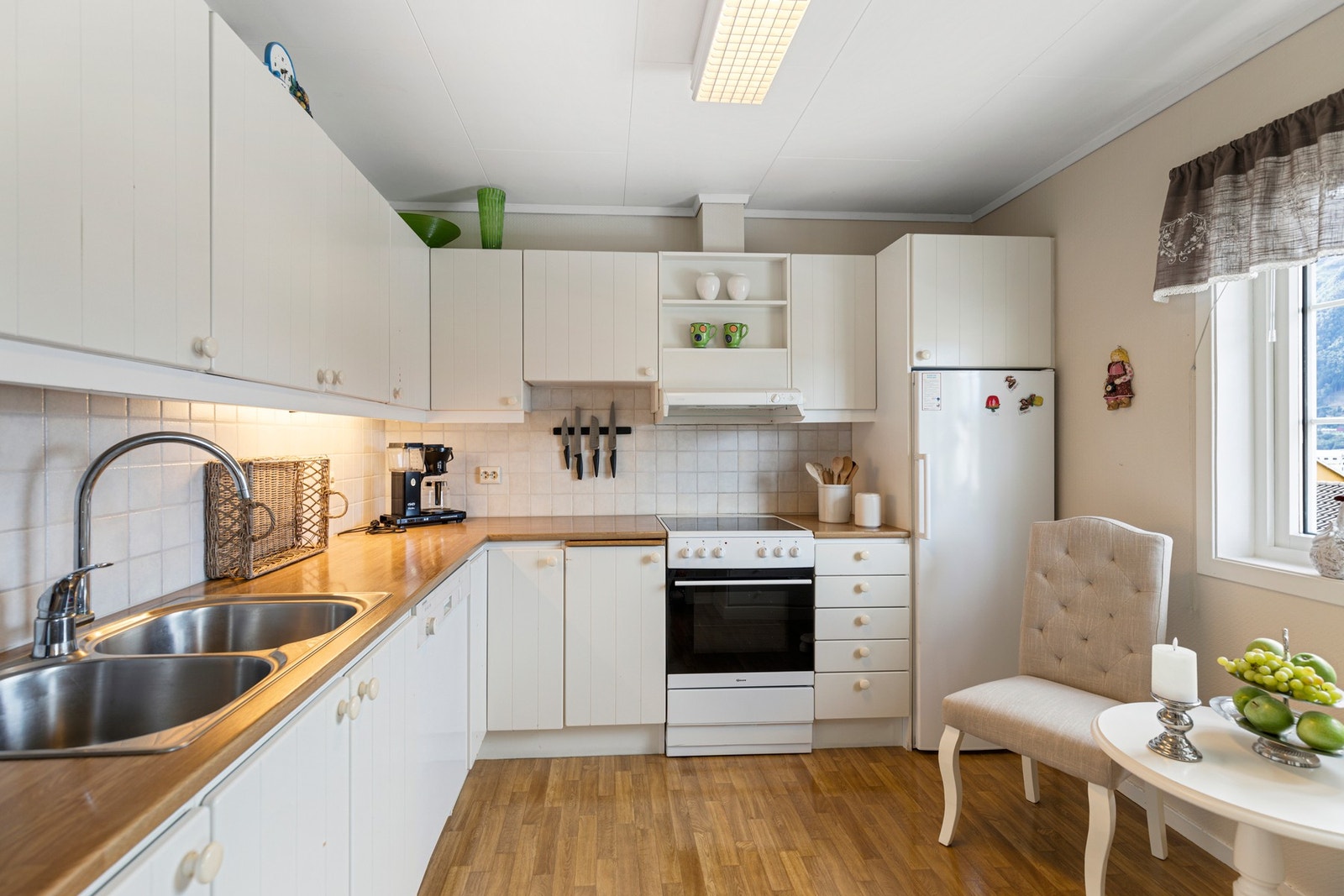 Fra stue er det tilkomst til kjøkkenet. Kjøkkenet har original innredning i hvite, profilerte fronter. . Innredningen inneholder videre laminat benkeplate med flis mot overskap, ventilator og opplegg for komfyr. Hvitevarer medfølger ved handel. Galleribilde