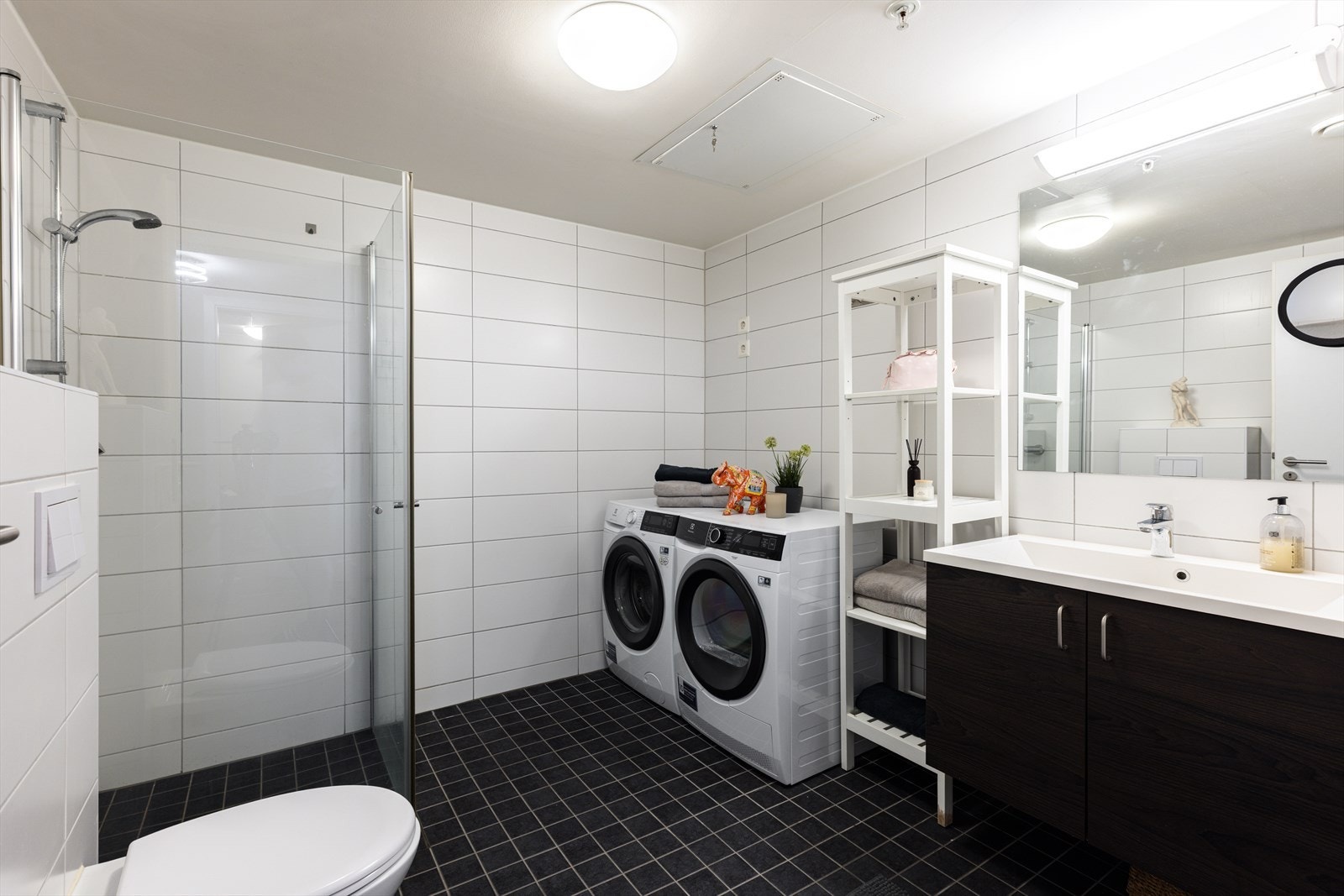 Pent baderom med fliser på gulv og vegg. Utstyrt med servant i skap, vegghengt toalett, dusjhjørne med innfellbare dører og opplegg for vaskemaskin. Rørfordelingsskap og stoppekran Galleribilde