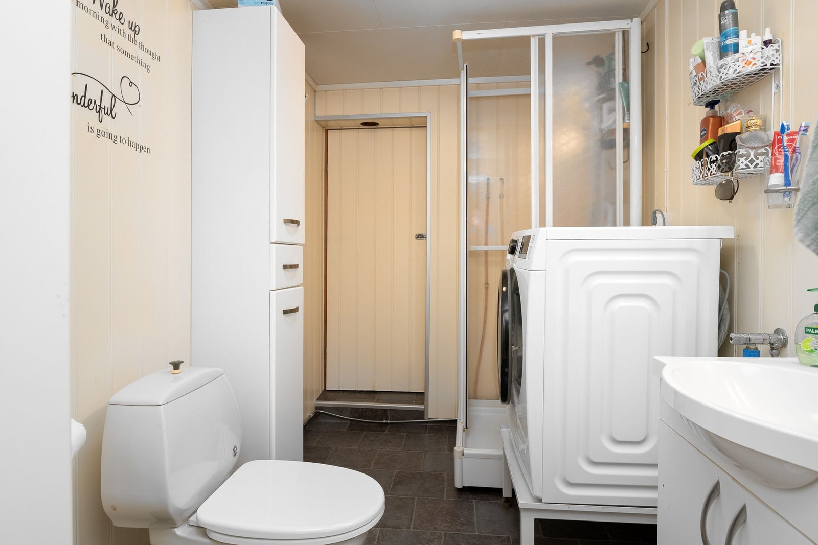 Bad/vaskerom utstyrt med servant på innredning, høyskap, toalett, dusjkabinett og opplegg for vaskemaskin. Galleribilde