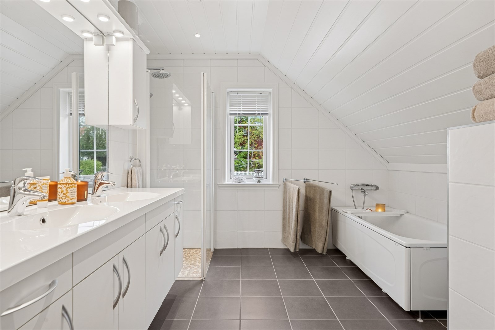 Badet er utstyrt med vegghengt toalett, dusjhjørne med glassdører, badekar, samt baderomsinnredning med to servanter og blandebatteri. Galleribilde