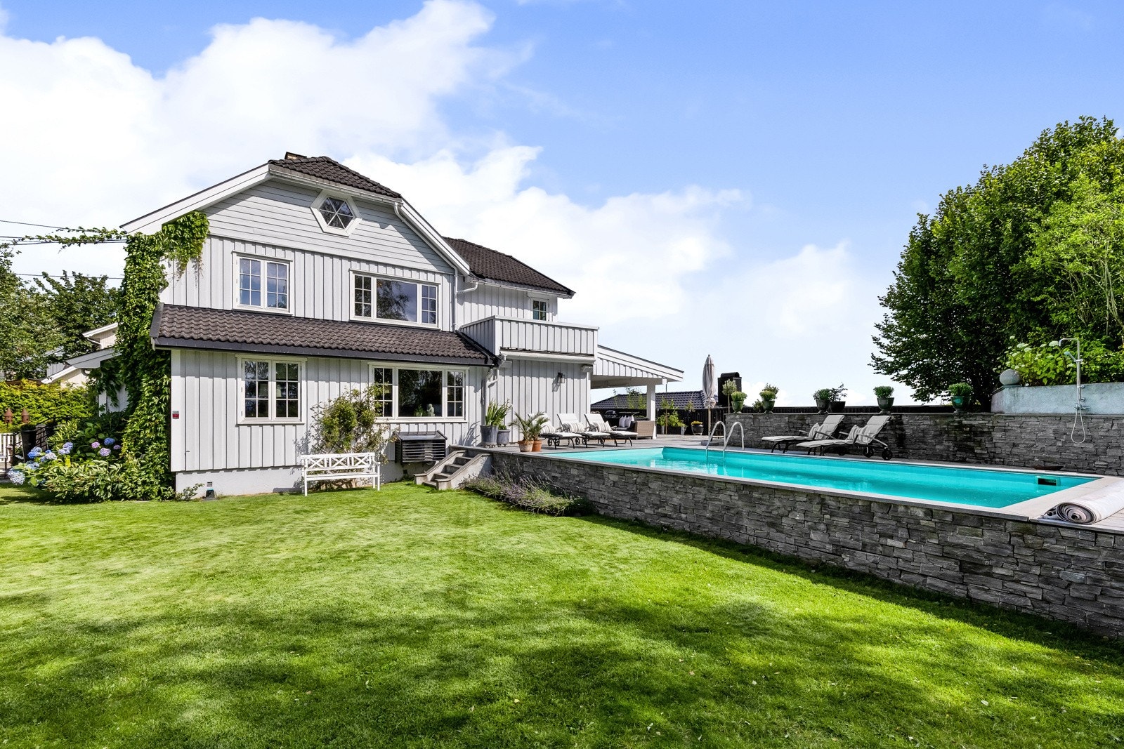 Terrassen rundt bassenget er som et deilig soldekk, skjermet av en dekorativ steinmur og trær på den ene siden, og med trapp ned til plenarealene i hagen.