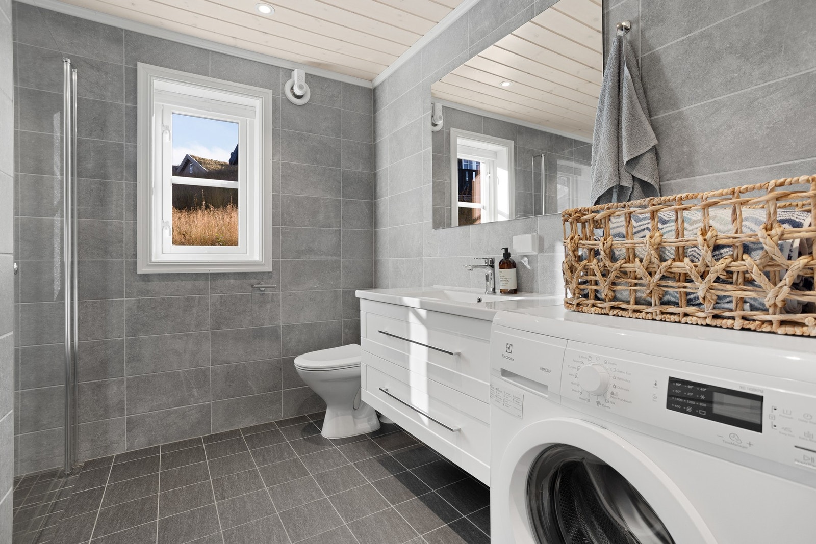 Baderom med flislagte gulv og veggflater. Baderommet inneholder toalett, innredning med servant, opplegg til vaskemaskin og dusjnisje med glassdør Galleribilde