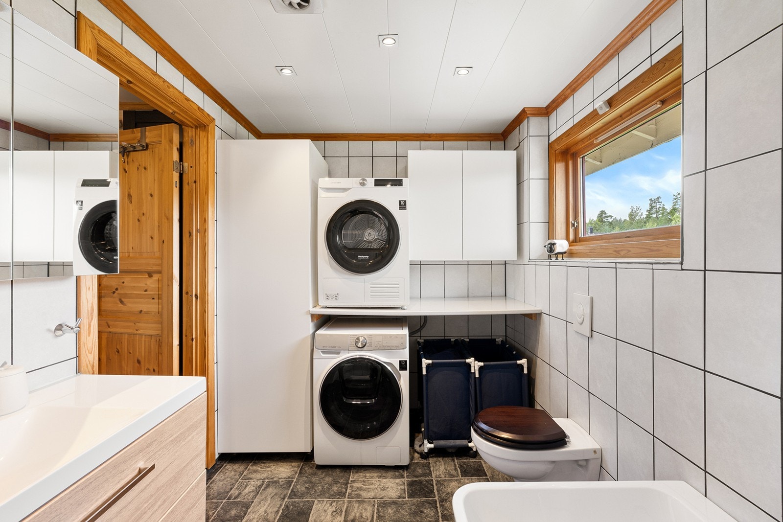 Rommet har en praktisk vaskesone med både vaskemaskin og tørketrommel i søyle, samt skap og benkeplass for klesvask. Galleribilde
