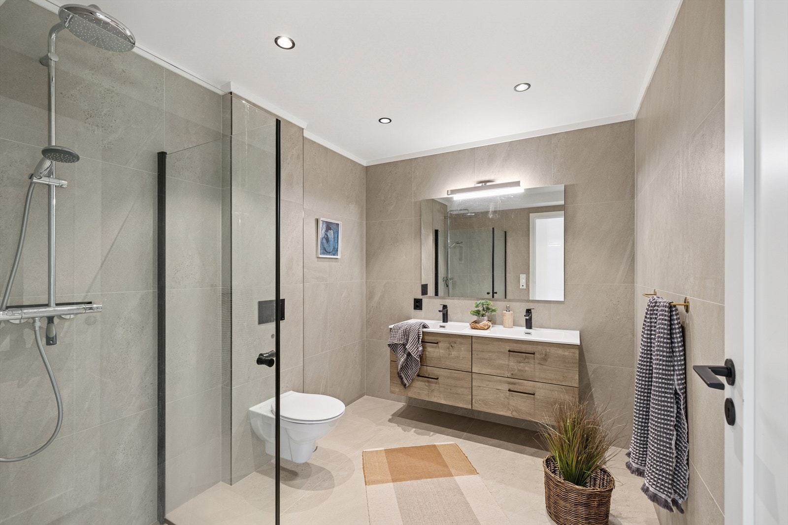 Bad med fliselagte overflater på vegger og gulv. Badet er innredet med baderomsinnredning med dobbel servant, speil med tilhørende belysning, dusjvegger og vegghengt toalett. Galleribilde