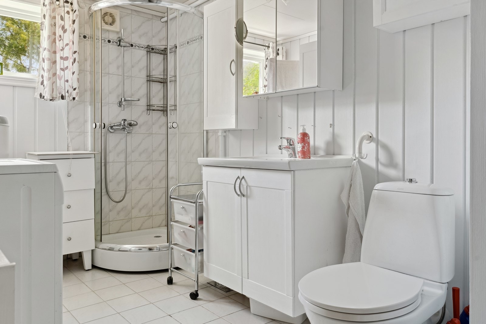 Bad med varmekabler og flis på golv og vegg i dusjsone, og malt panel på veggar. Det er dusjkabinett, skapinnreiing med heilstøypt servant, speilskap og golvståande toalett. Det er også godt med plass for oppbevaringsmøblar og vaskemaskin Galleribilde