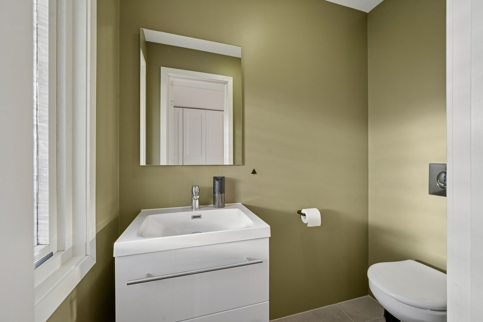 Det er et eget praktisk toalettrom i hovedetasjen, som er moderne gjennomført i moderne farger på veggene og flislagt gulv. Rommet er innredet med vegghengt toalett og baderomsinnredning med nedfelt servant, i tillegg til vegghengt speil. Galleribilde