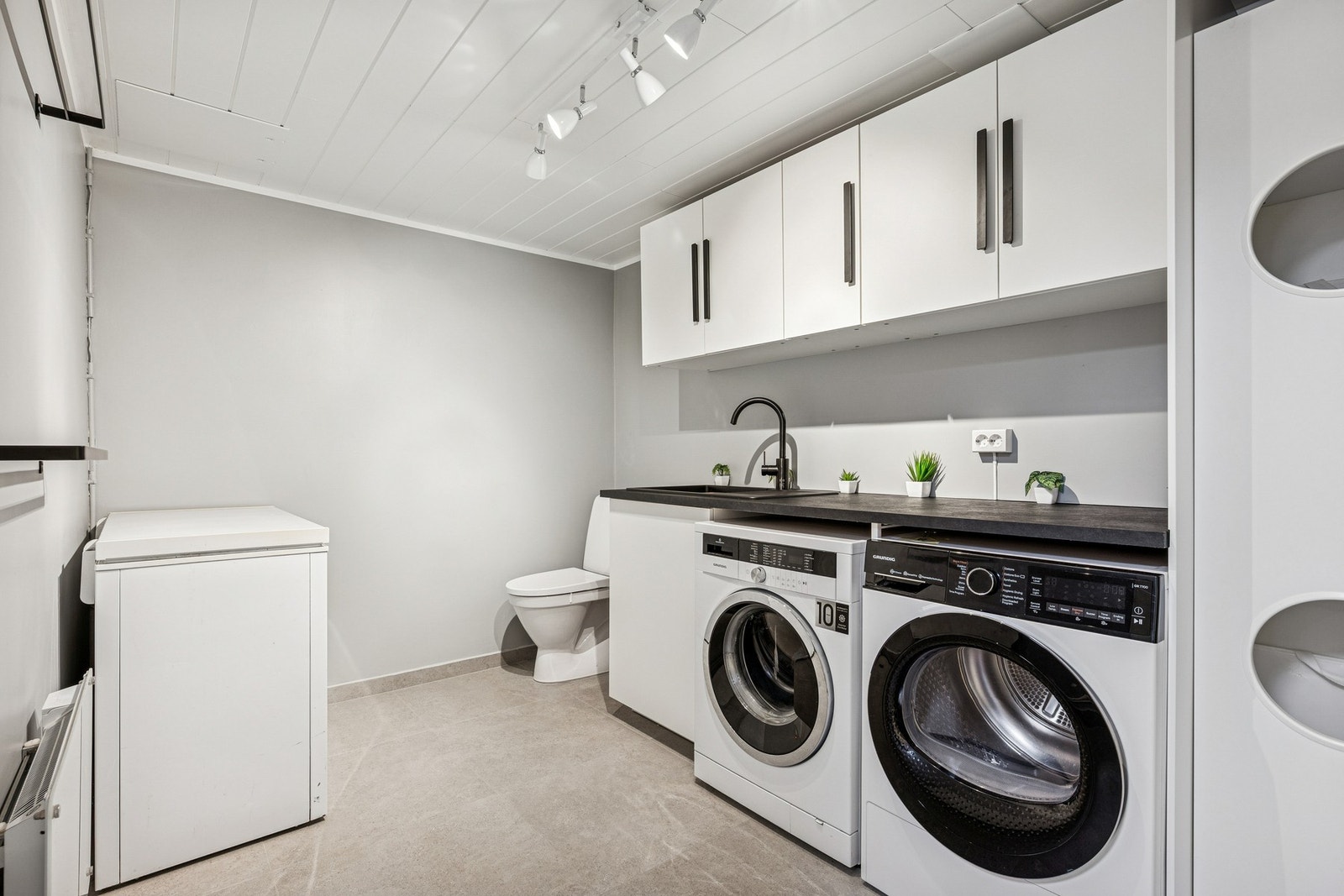 Moderne toalett- og vaskerom med flislagt gulv, malte vegger og stilren innredning med laminert benkeplate. Rommet har plass til vaskemaskin og tørketrommel. Galleribilde