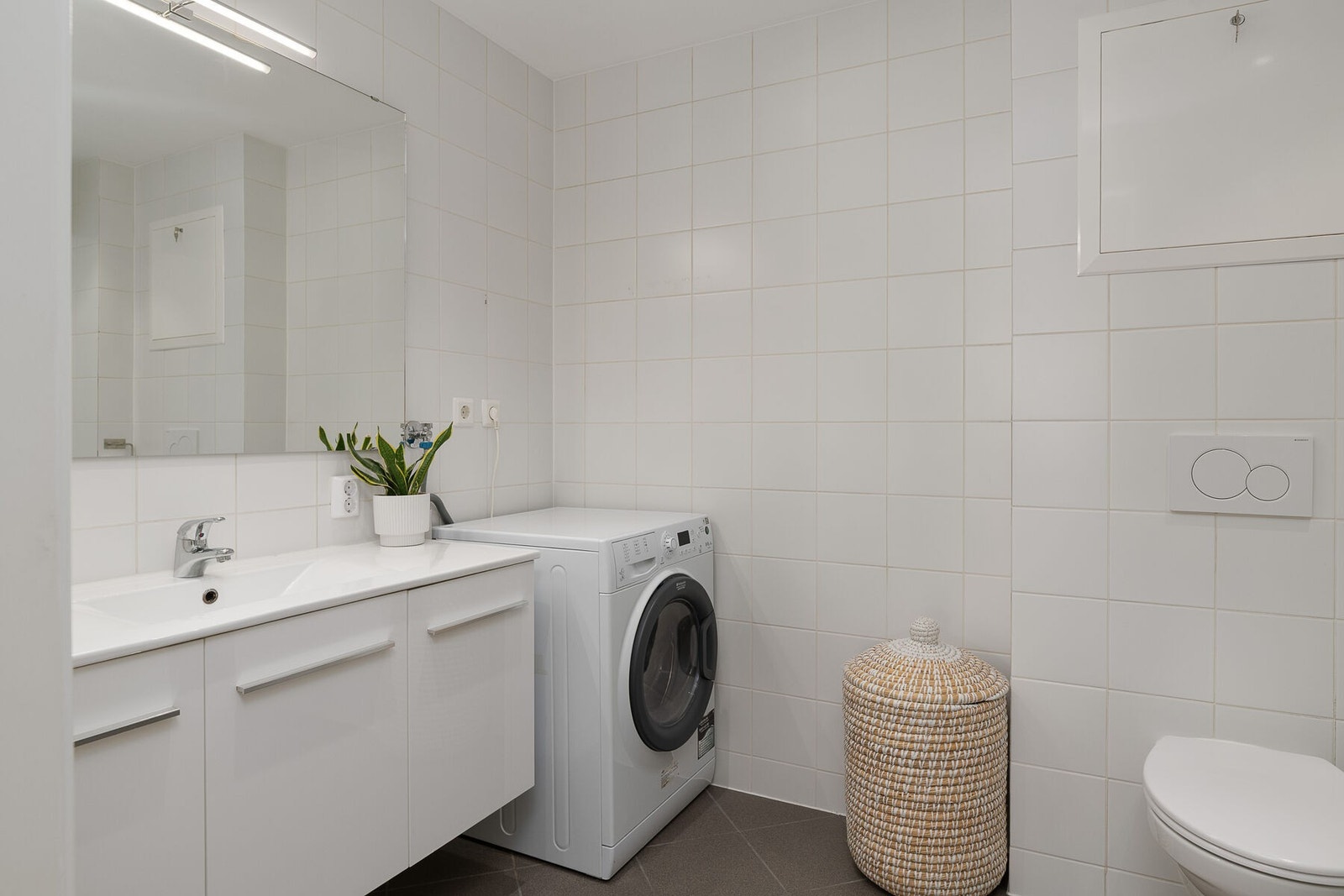 Flislagt bad/vaskerom i tidløs utførelse. Badet har servant på innredning, høyskap, veggmontert toalett, dusjkabinett og opplegg for vaskemaskin. Galleribilde