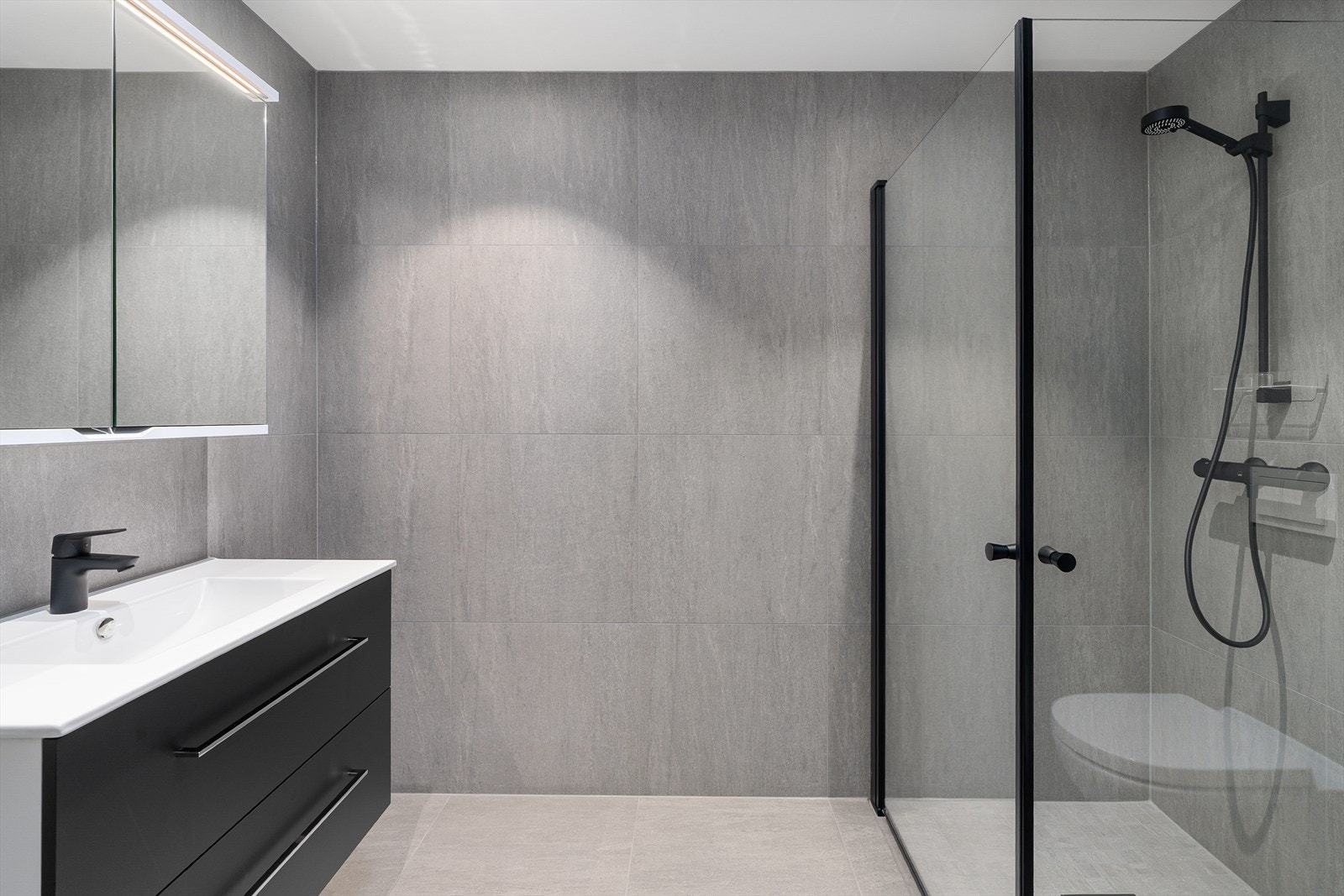 Lekkert bad med fliser på gulv og vegger. Utstyrt med vegghengt toalett, dusj, samt baderomsinnredning med heldekkende servant, speil med skap og god belysning. Galleribilde