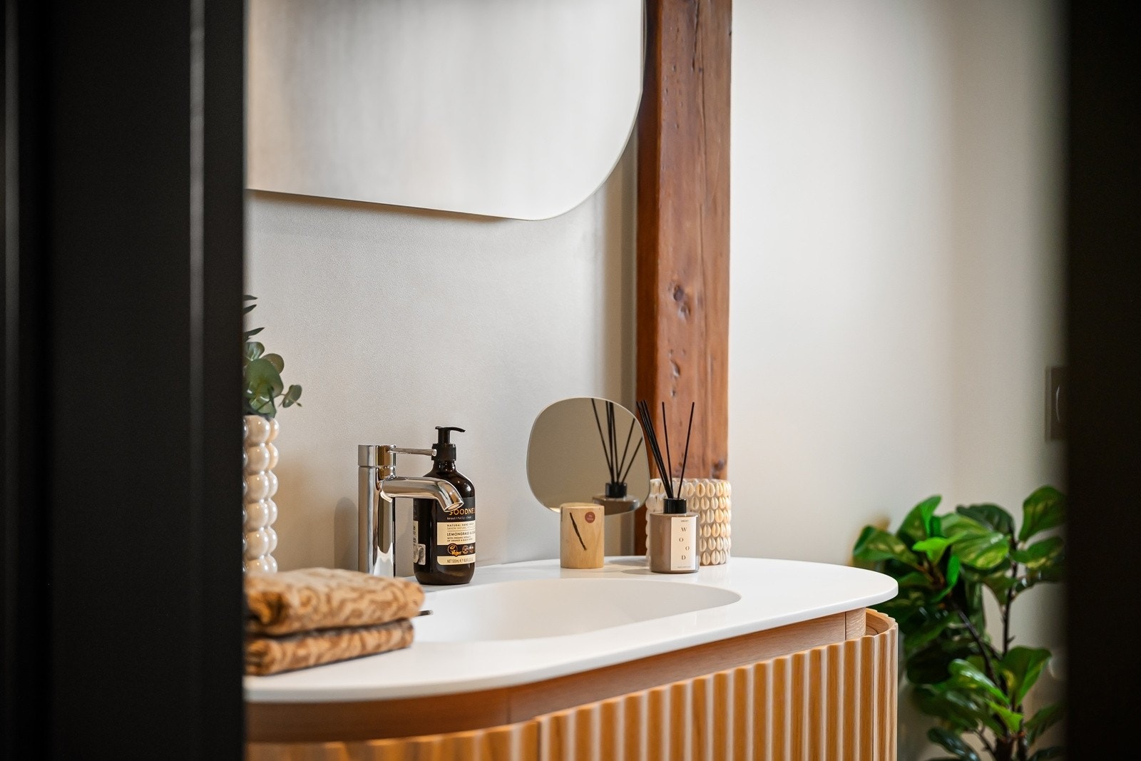 Oval corianservant på servantmøbel i eik med buede rillefronter, et matchende ovalt speil med indirekte belysning og et veggmontert toalett. Galleribilde