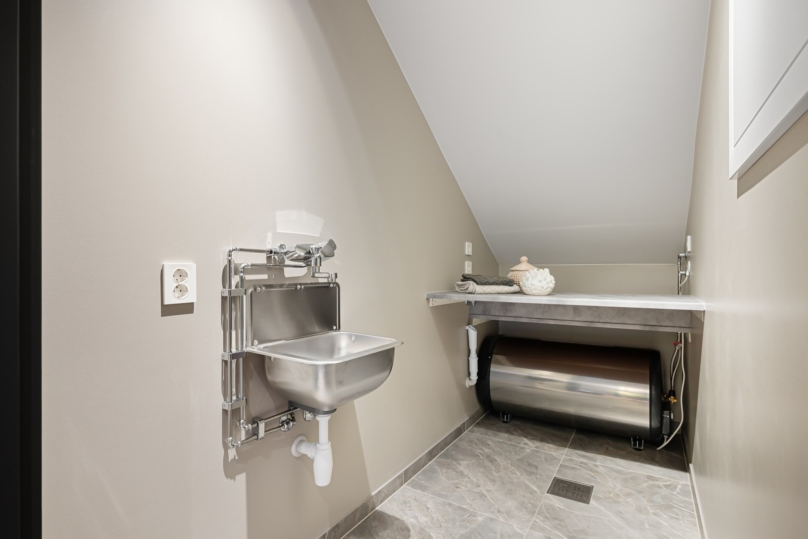 Svært praktisk vaskerom med flislagt gulv og gulvarme, utslagsvask og nisje for vaskemaskin og tørketrommel under benkeplate. Galleribilde