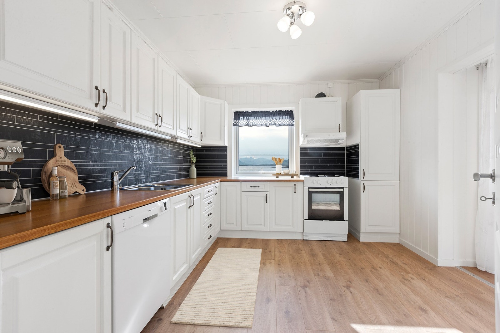 Kjøkken med lyse overflater, praktiske oppbevaringsløsninger og benkeplass, samt nydelig utsikt. Galleribilde