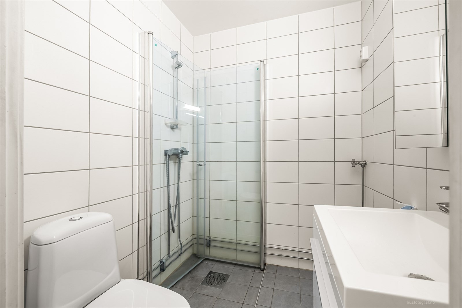 Pent flislagt bad med innredning bestående av klosett, dusjhjørne og baderomsinnredning med speilskap. Galleribilde