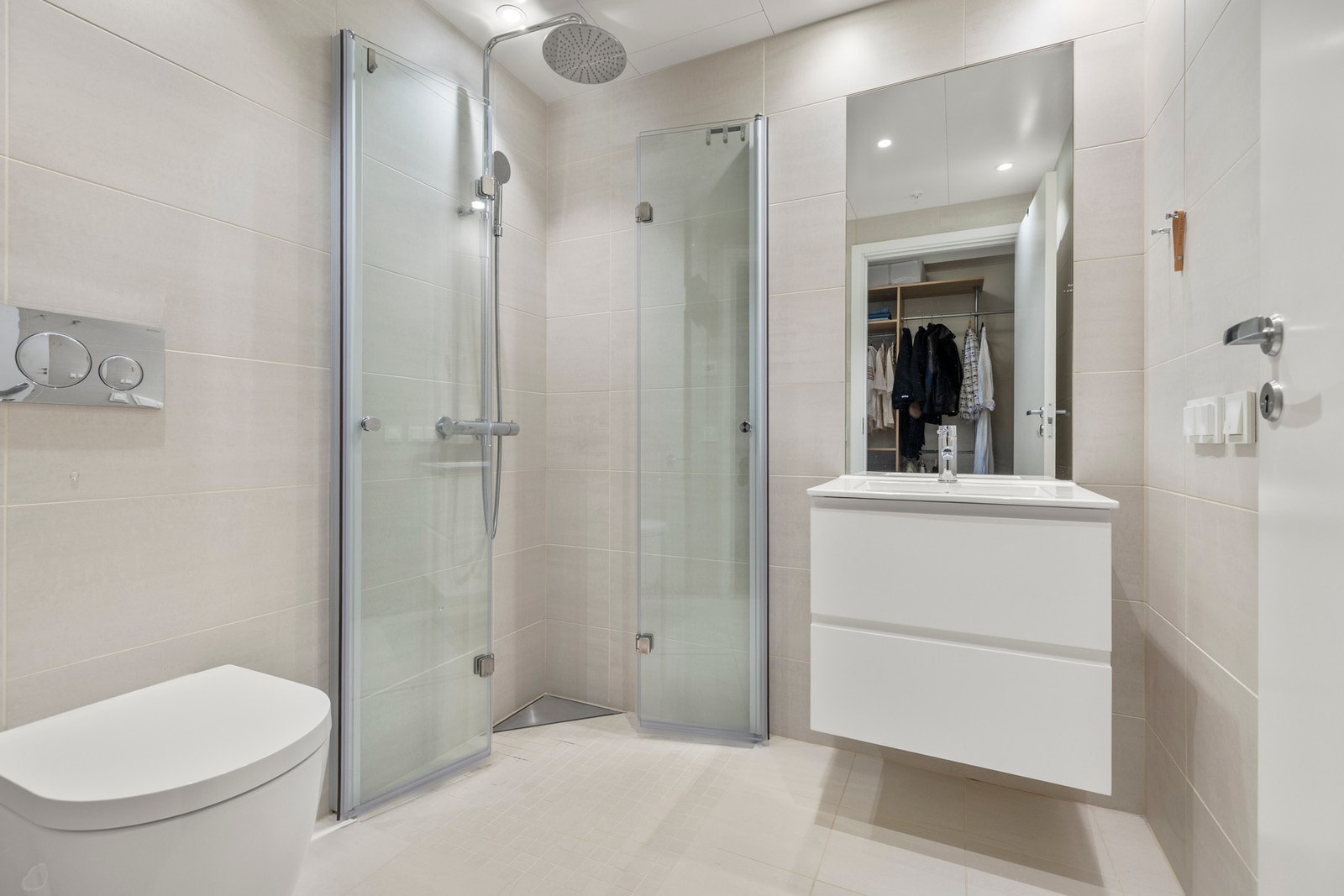 Badet har flislagte overflater, himlingsplater med downlights, servant med skuffer, speil, vegghengt toalett og dusjhjørne med to foldbare glassdører. Galleribilde