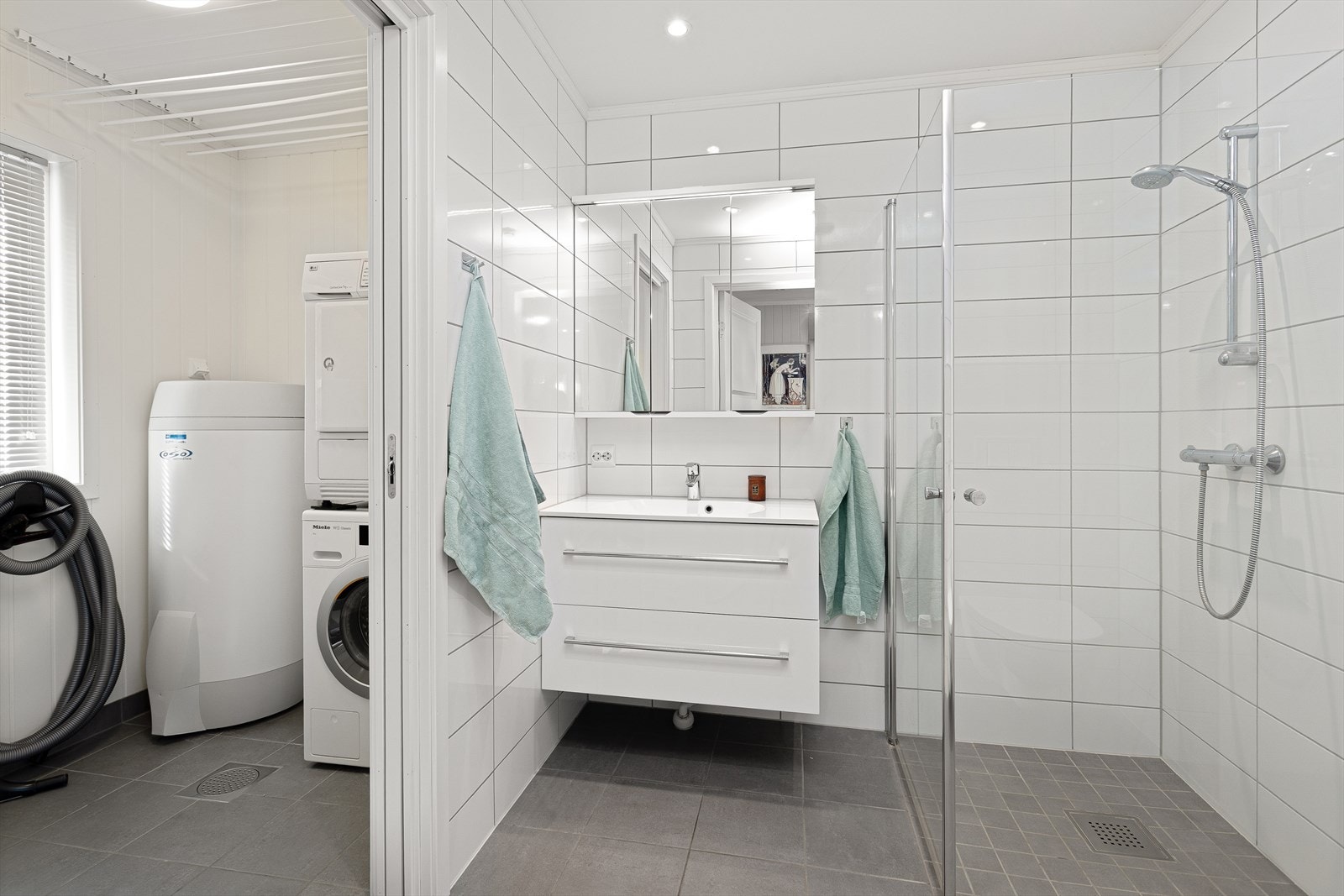 Badet er utstyrt med dusjhjørne, vegghengt wc og en moderne baderomsinnredning. Videre inngang til vaskeroms-seksjon. Galleribilde