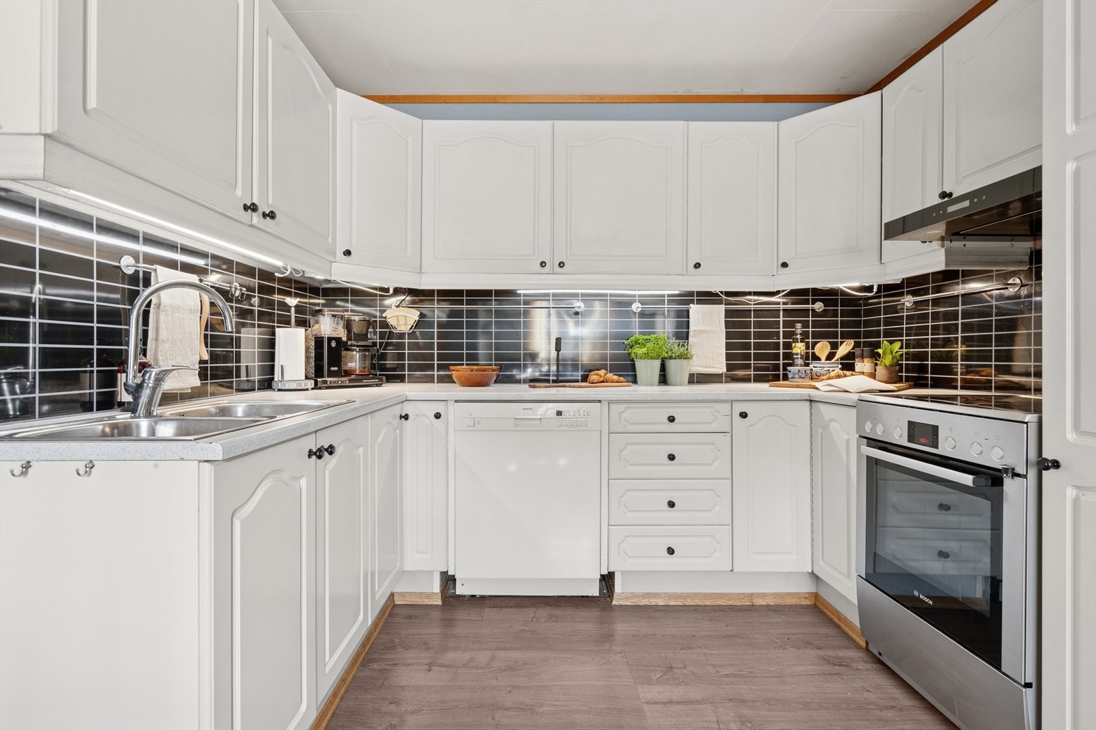 Kjøkkenets innredning har malte, profilerte fronter, laminert benkeplate, kitchenboard på vegg over benk, dobbel stålvask og belysning under overskap. Galleribilde