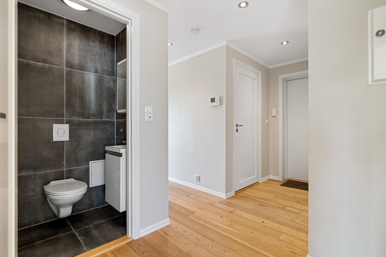 Rommet er innredet med vegghengt toalett, elegant servantskap og speilskap som gir både oppbevaringsplass og stil. Elektriske varmekabler gir behagelig gulvvarme, og godt inneklima sikres med mekanisk avtrekk i taket og tilluft via døren. Galleribilde