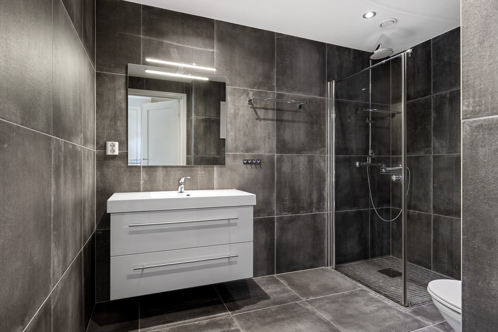 Badet fremstår lekkert og innbydende, med fliser på både gulv og vegger som gir et eksklusivt preg. Himlingen er elegant utført med malte plater og integrerte downlights som gir behagelig belysning. Galleribilde