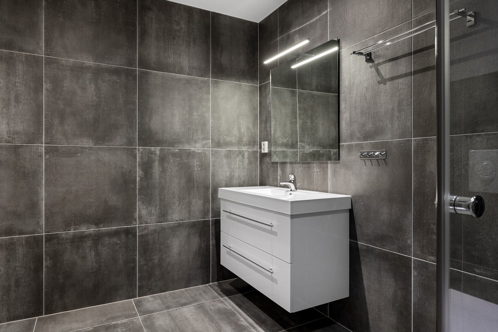 Rommet er funksjonelt innredet med et stilrent dusjhjørne, moderne servantskap med speil, vegghengt toalett og opplegg for vaskemaskin. Elektriske varmekabler sørger for deilig gulvvarme. Galleribilde