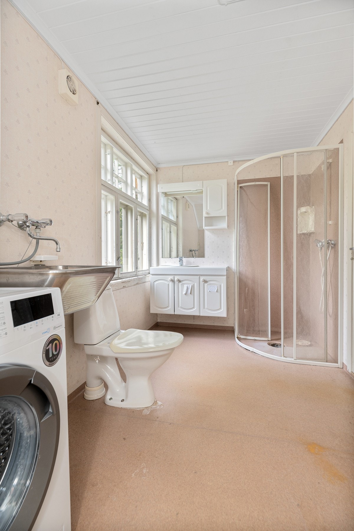 Badet er eldre med gulvbelegg, lys servantinnredning, toalett og dusjhjørne med skyvedører.
Vaskemaskin medfølger. Galleribilde