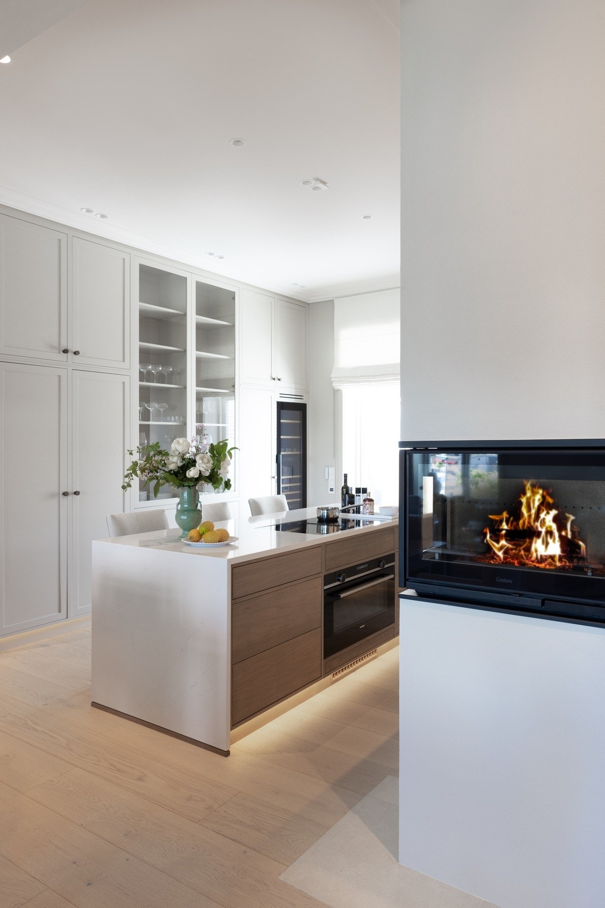 Kjøkken med fronter i en kombinasjon av eik og malt MDF, benkeplate i steinkompositt og integrerte hvitevarer fra Siemens. Praktisk kjøkkenøy, peis og høyt vinskap gir en elegant og helhetlig stil. Galleribilde