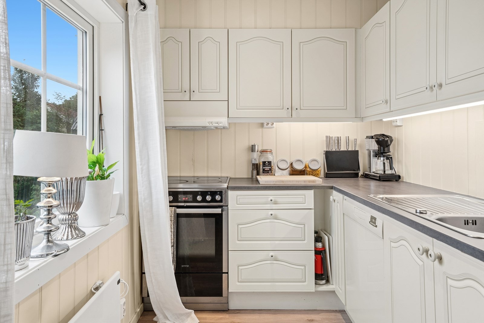 Kjøkkeninnredning med profilerte fronter og laminat benkeplate med nedfelt vaskekum. Kjøkkenet er utstyrt med belysning over benkeplaten, overskap og kjøkkenventilator med avtrekk ut. Galleribilde