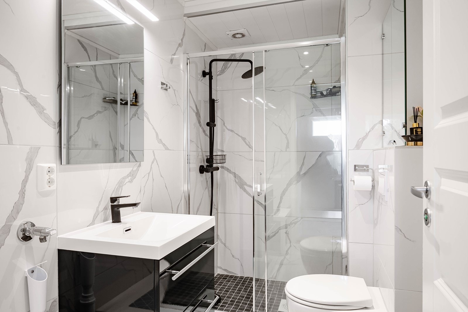 Moderne baderomsinnredning med skuffer og heldekkende servant, speil med belysning over. Badrommet er også utstyrt med romslig dusj med skyvedør av gladd og både hånd- og regnfallsdusj, speilskap og vegghengt toalett. Galleribilde