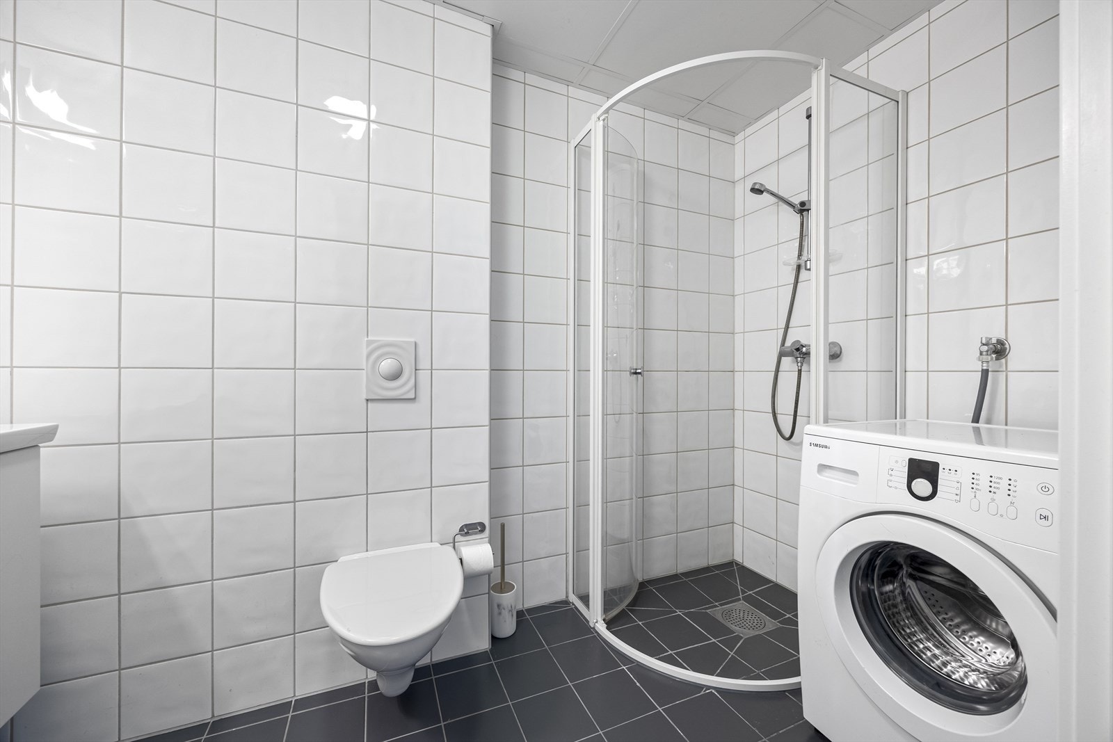 Fliselagt bad med vegghengt toalett, dusjhjørne og opplegg for vaskemaskin. Galleribilde