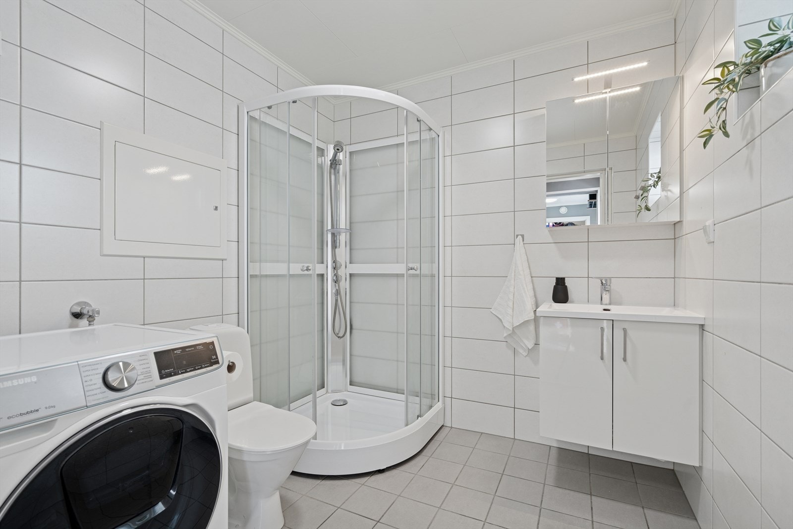 Badet er utført med grå gulvfliser m/varmekabler under og hvite avlange veggfliser - Her finner du toalett, dusjkabinett, opplegg for vaskemaskin og baderomsinnredning Galleribilde