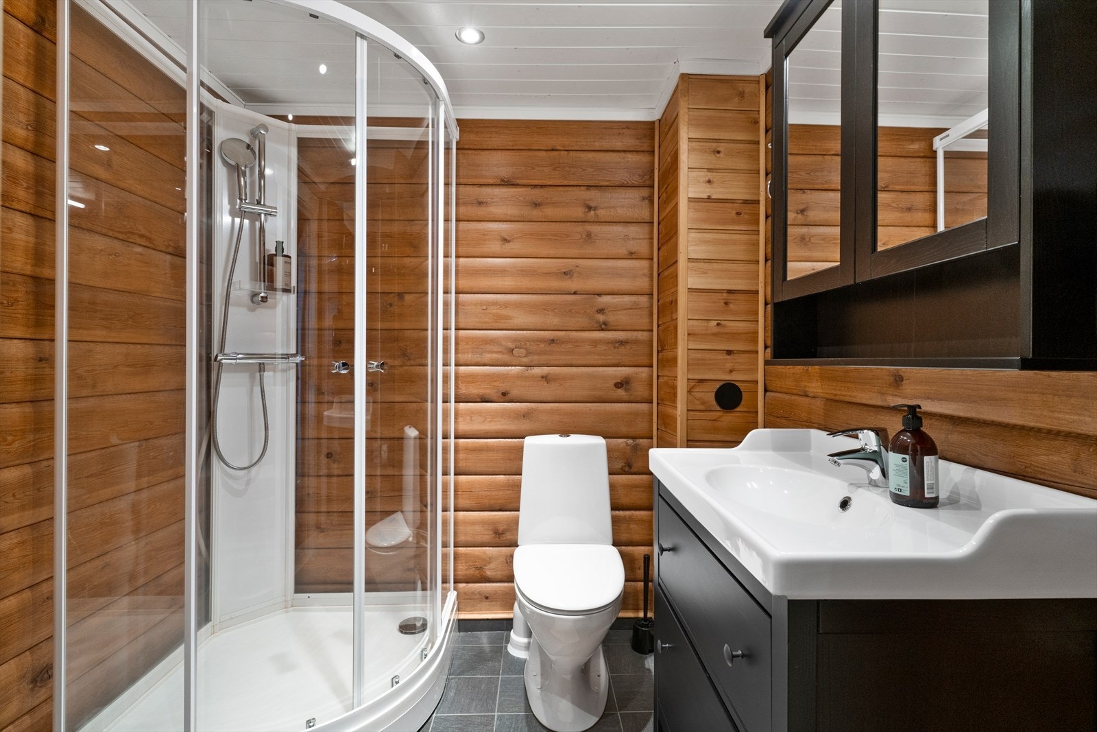 Badet inneholder behandlet innredning med servanttopp, skap på vegg med speil og dører montert over servant, toalett, dusjkabinett og opplegg for vaskemaskin. Galleribilde