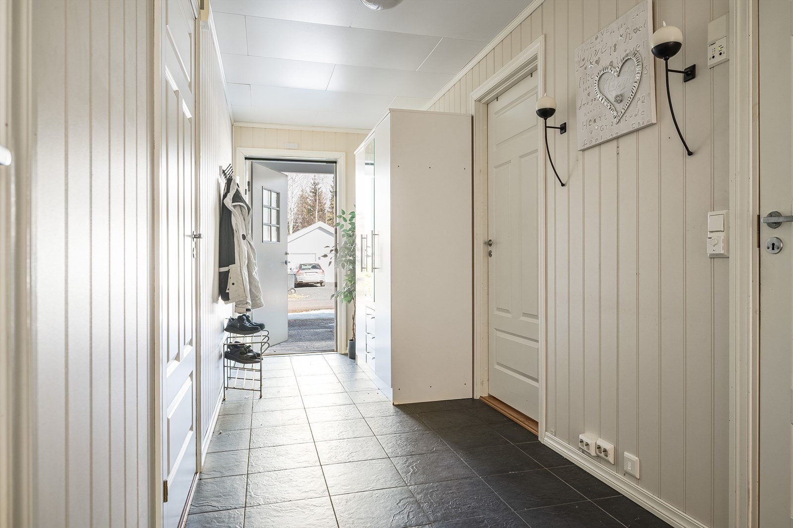 Du ønskes velkommen inn i en lys, praktisk og romslig entré med plass til sko og yttertøy i medfølgende garderobeskap. Entréen har mørke fliser på gulv mee gulvvarme, mate panelplater på veggene og skjult elektrisk anlegg. Galleribilde