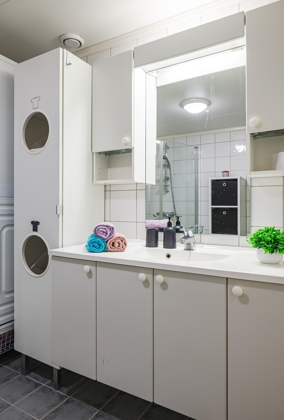 Lys baderomsinnredning med heldekkende servant, speil med belysning og vegghengte skap over. I hjørnet er det plass og opplegg for vaskemaskin og tørketrommel over om ønskelig. Galleribilde