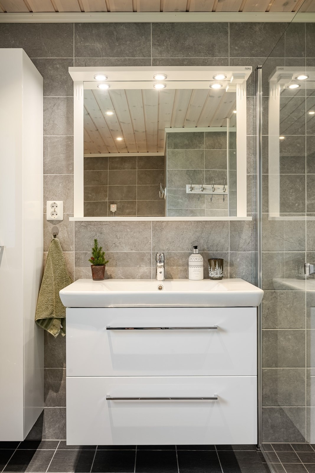 Moderne baderomsinnredning med skuffer og heldekkende servant, speil med belysning over, samt ekstra høyskap ved siden av. Galleribilde