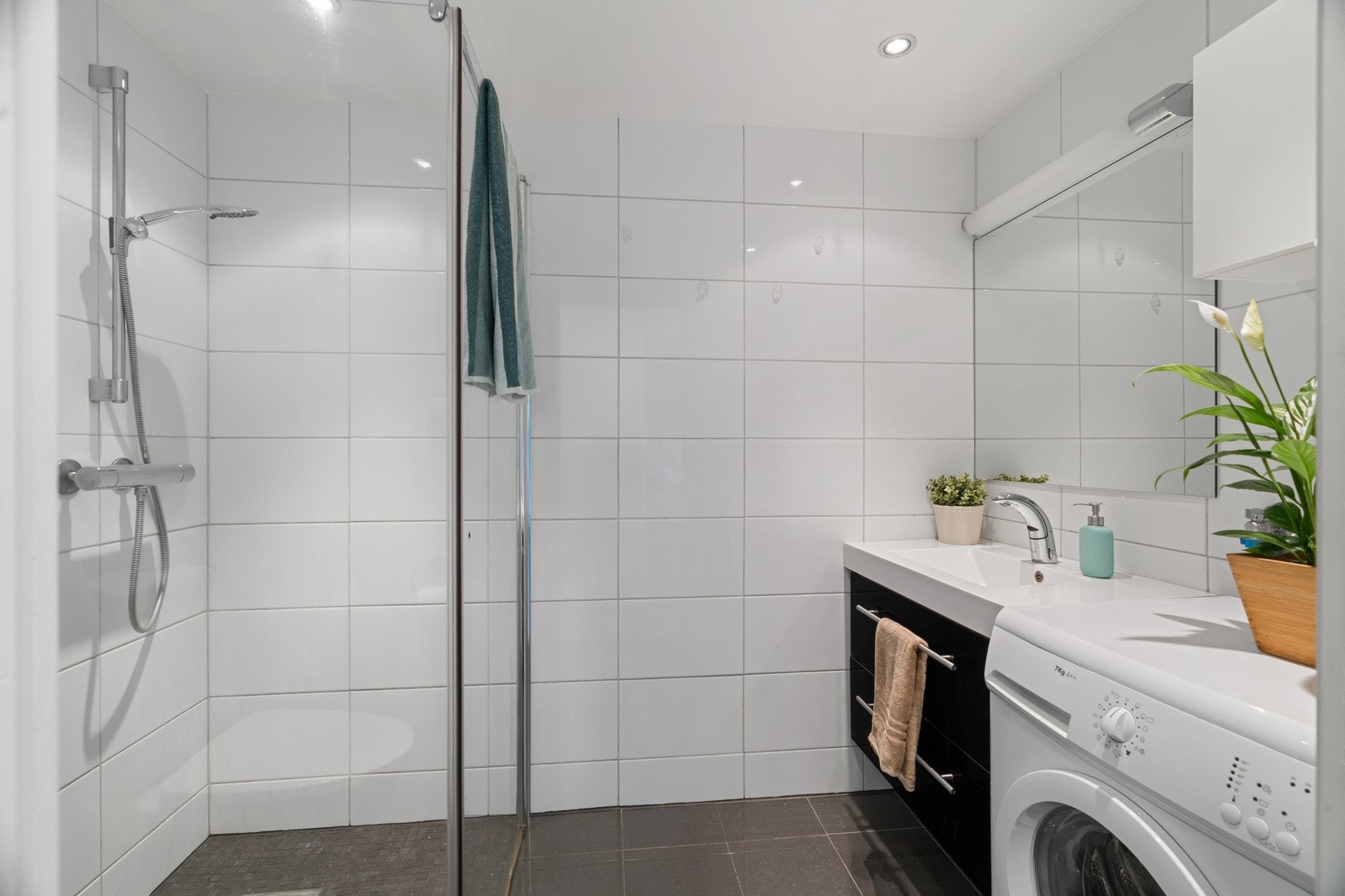 Flislagt baderom med varmekabler i gulv og praktiske dusjvegger. Badet har en servant, med en mørk innredning med speil, downlights i himling samt opplegg for vaskemaskin. Galleribilde
