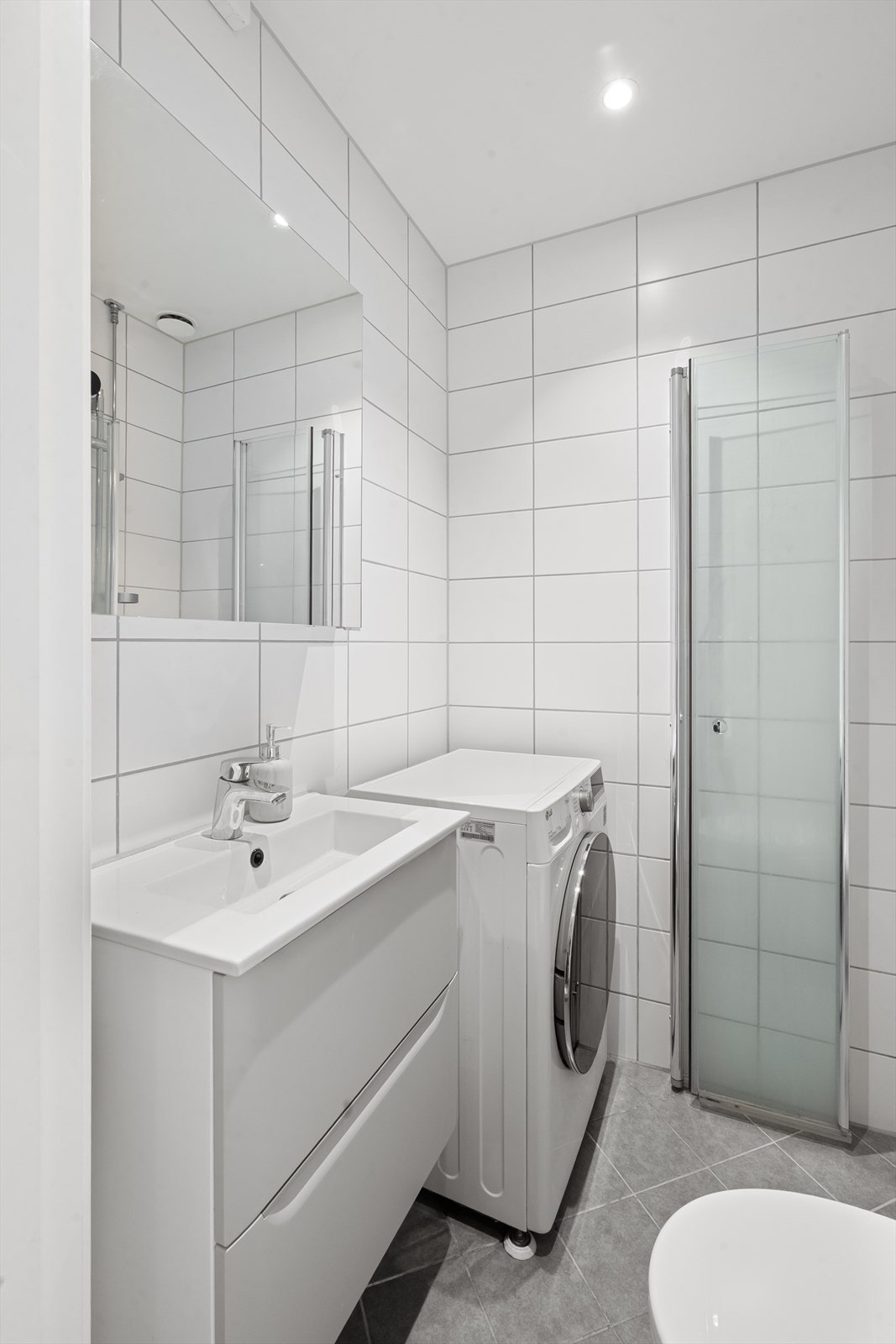 Badet har opplegg for vaskemaskin, servant med skuffer, speilskap, toalett og hjørnedusj med innsvingbare dører i glass. Galleribilde